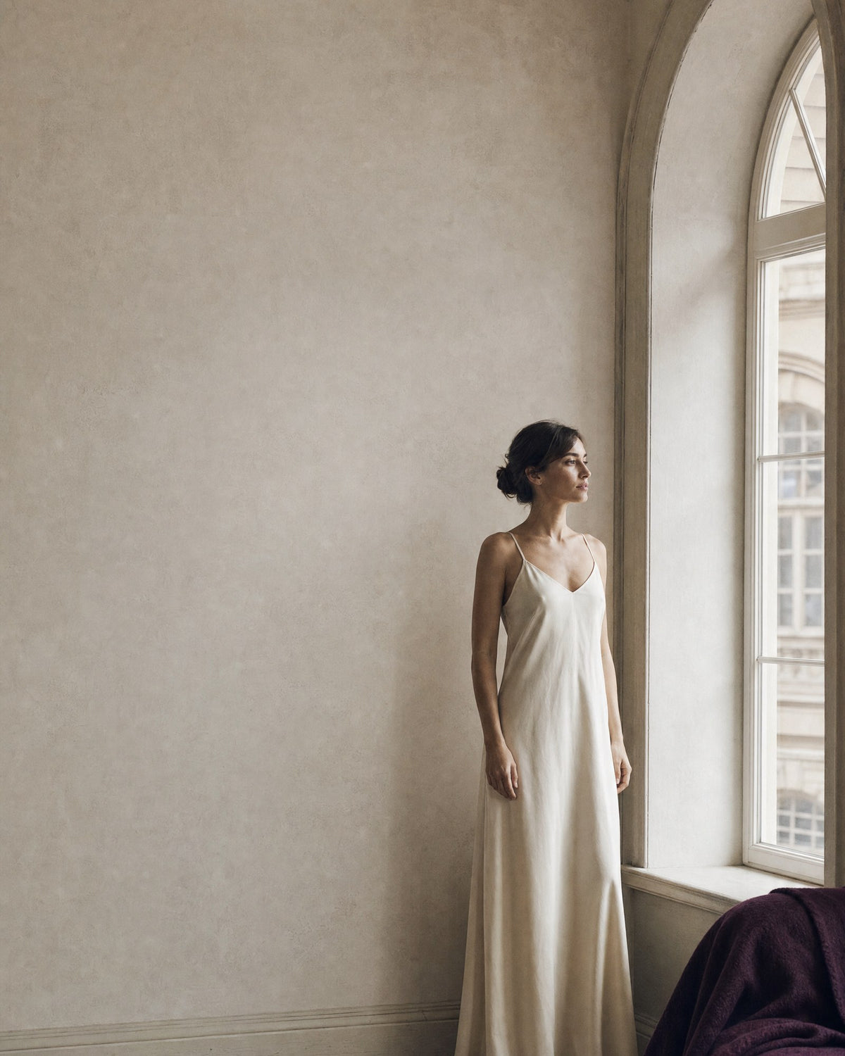 Woman in a floor length cream slip dress standing beside an arched window